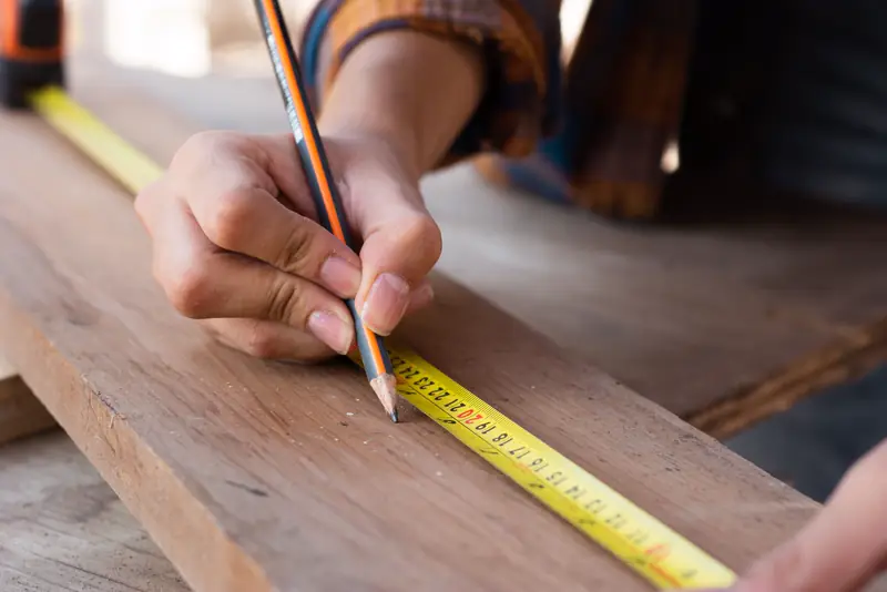 Hands marking a cut line on a board with a pencil and a yellow tape measure